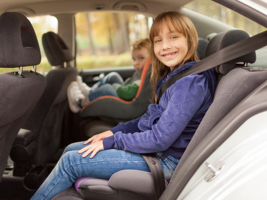 A girl in a booster seat wearing a seat belt, with a younger child in a car seat behind her