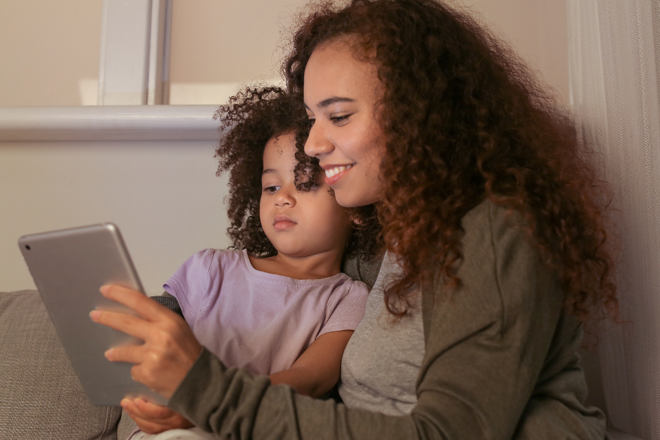 A mother and her young daughter looking at a tablet together on a sofa