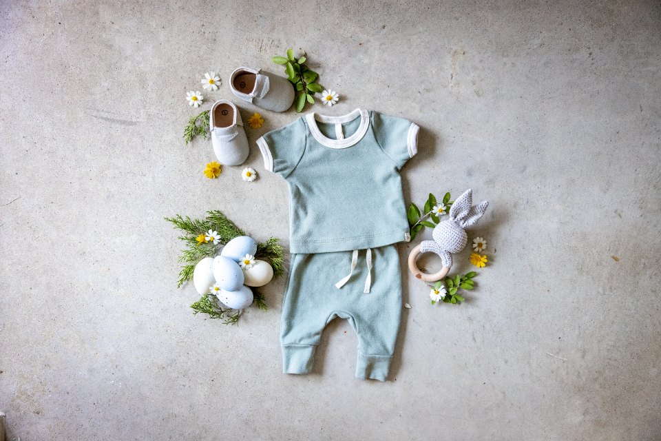 Baby clothes, small shoes and a crocheted rattle laid out on a concrete floor